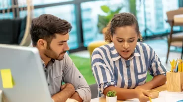 Two people discussing a project at a desk with documents and a small plant.