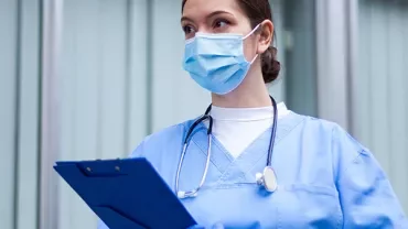 Healthcare worker in blue scrubs holding a clipboard and wearing a mask.