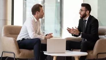 Two people sitting in an office setting, engaged in a discussion with a laptop on the table.