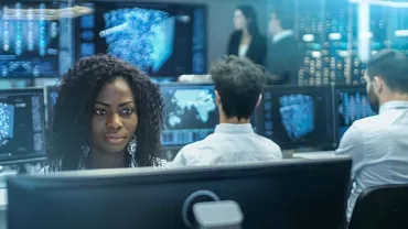 People working at computers in a tech office, multiple screens showing data visualizations.