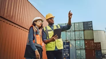 Two workers in safety gear at a shipping yard, one pointing while holding a tablet.
