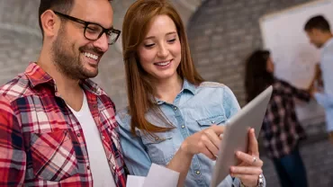 Two people looking at a tablet, smiling, in a casual office setting.