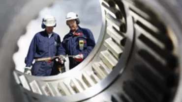 Two workers inspecting large machinery gears, wearing protective gear.