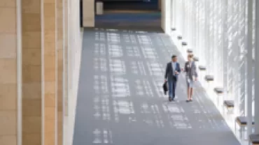 Two people walking in a bright, modern hallway with large windows.