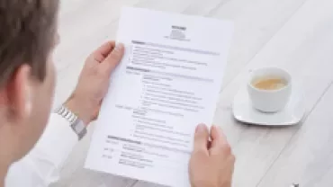 Person holding a printed resume at a desk with a cup of coffee.