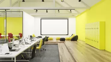 Modern office space with yellow walls and lockers, featuring desks and a projector screen.