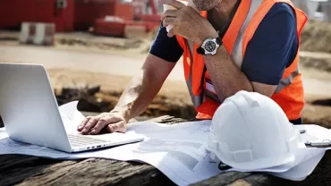 Construction worker using a laptop on site with blueprints and a hard hat nearby.