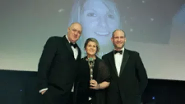 Three people in formal attire pose on stage with an award.