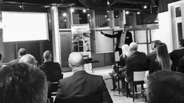 A presenter pointing at a screen in front of an audience in a modern conference room.