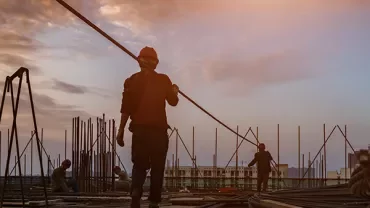Construction workers on a building site at sunset, carrying and positioning steel rods.